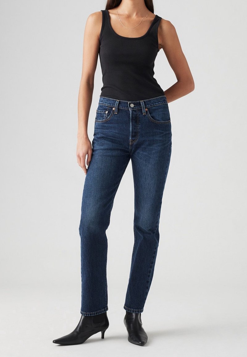 Jean en denim bleu foncé avec une coupe droite, doté de cinq poches et d'une fermeture éclair. Associé à un débardeur noir ajusté et des bottes à talons noires.