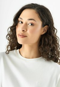 Weißer Sweatshirt mit Rundhalsausschnitt und dezentem, strukturiertem Blumenstickerei auf der Vorderseite. Das Model hat lockige Haare und ein natürliches Make-up.