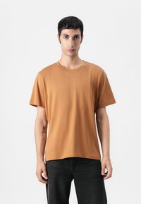 T-shirt à col rond à manches courtes en tissu de coton orange rouille, texture lisse, coupe décontractée, associé à un jean noir, le modèle se tient devant un fond uni.