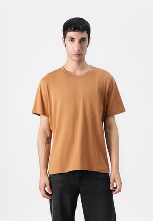 Kortærmet t-shirt med rund hals i rust-orange bomuldsstof, glat tekstur, afslappet pasform, parret med mørke jeans, model står foran en ensfarvet baggrund.