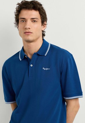 Joven de pie vestido con una camiseta polo azul, pantalones beige claros y zapatillas blancas sobre un fondo liso y claro.