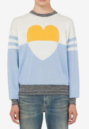Maglione con motivo a cuore giallo e bianco su sfondo blu e bianco, collo, polsini e orlo a coste grigio, indossato con jeans in denim.