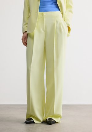 Osoba ubrana w jasnopsoilone spodnie o szerokich nogawkach, dopasowaną marynarkę, niebieską bluzkę oraz czarne buty na szpic z złotymi klamrami.