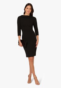 Kobieta w dopasowanej czarnej sukience do kolan z rękawami do łokci, trzymająca czarną kopertówkę i nosząca beżowe szpilki na kostkę.