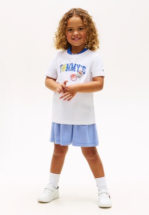 Παιδί με σγουρά μαλλιά που φοράει λευκό μπλουζάκι με σχέδιο, μπλε και άσπρη ριγέ φούστα, λευκές κάλτσες και λευκά αθλητικά παπούτσια, στέκεται με τα χέρια ενωμένα.