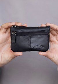 Černá kožená peněženka na zip, která má přední kapsu, prošité detaily a embosované logo. Kompaktní a obdélníkový tvar.