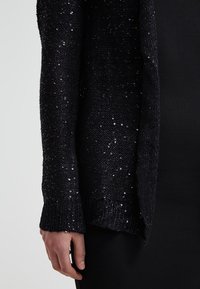 Svart stickad cardigan dekorerad med paljetter, med en texturerad yta, öppen fram och ribbade muddarna, som visar glans och intrikata detaljer.