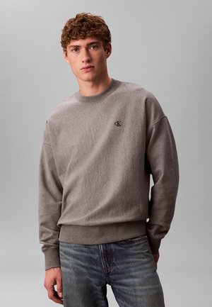 Młody mężczyzna z kręconymi włosami, ubrany w szarą bluzę Calvin Klein i niebieskie dżinsy, stojący na jednolitym szarym tle.