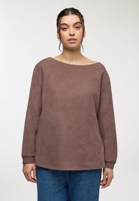 Brązowy, teksturowany sweter z długim rękawem, z szerokim dekoltem, luźnym krojem i ściągaczami, zestawiony z niebieskimi dżinsami.