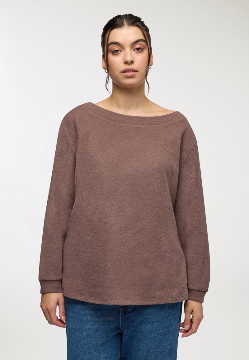 Brązowy, teksturowany sweter z długim rękawem, z szerokim dekoltem, luźnym krojem i ściągaczami, zestawiony z niebieskimi dżinsami.