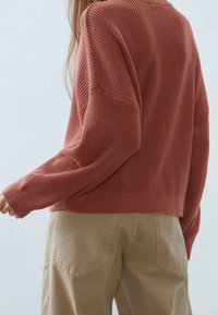 Rosenroter Strickpullover mit lockerem Schnitt, ausgestattet mit gerippten Bündchen und Saum, kombiniert mit hellbeigen Hosen.