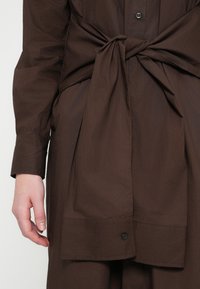 Καφέ πουκάμισο με κουμπιά μπροστά και λεπτομέρεια δεσίματος στη μέση, με μακριά μανίκια και ομαλή, υφή υφάσματος. Περιλαμβάνει κούμπωμα με ένα κουμπί στο κάτω μέρος.