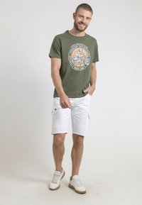 T-shirt en coton vert avec graphique moto orange, associé à un short cargo blanc. Le modèle porte des baskets blanches et marron, debout de manière décontractée.