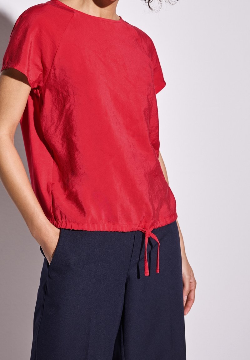 Röd kortärmad topp med dragsko i nederkant, tillverkad av ett mjukt och lätt tyg, bärs med mörkblå byxor.