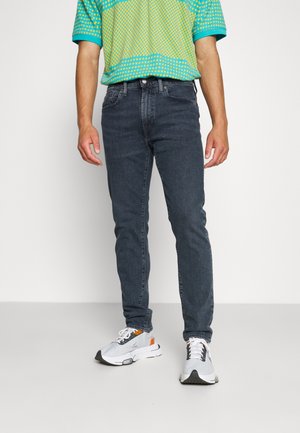 Homme portant un jean slim gris foncé, des baskets blanches avec des accents noirs et orange, et une chemise à manches courtes turquoise et jaune à motifs.