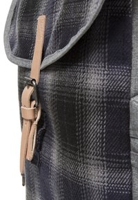 Närbild av en mörkgra och svart rutigt tyg ryggsäck med ett beige läderlåsband och svart metallspänne.