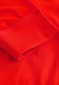 Gros plan d'un poignet de manche en tissu rouge avec une texture côtelée et des coutures visibles sur un fond lisse de vêtement rouge.