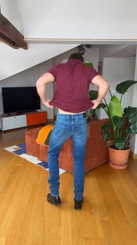 Oseba dviguje bordo majico, stoječa v dnevni sobi z lesenim tlemi, oranžnim kavčem, lončnico in televizorjem na nizki omarici.