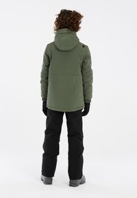 Veste isolante verte avec capuche, poignets élastiques et ourlet arrondi, associée à un pantalon noir imperméable et des bottes grises.