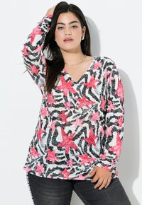 Vrouw met lang donker haar, die zilveren oorbellen en een ketting draagt, poseert in een lang mouw V-hals top met een roze ster en zwarte zebra strepen.