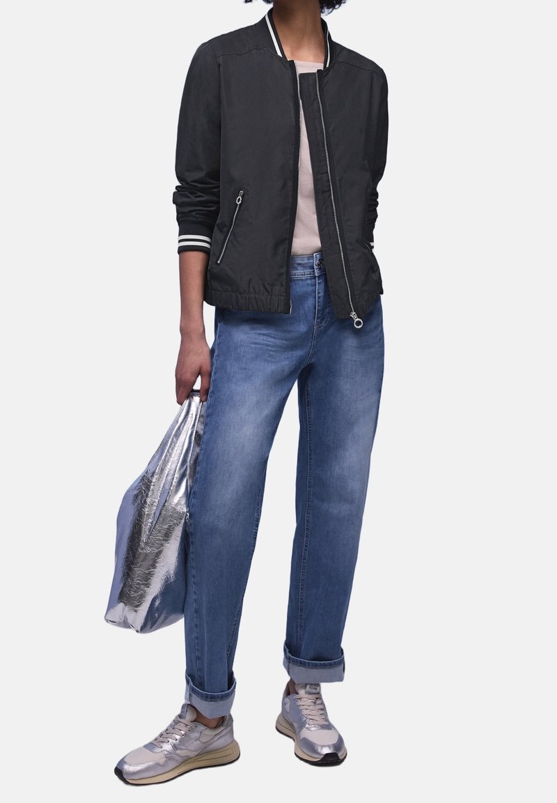 Personne portant une veste bomber noire, chemise rose clair, jean bleu retroussé, baskets argentées, tenant un grand sac métallique argenté à côté.