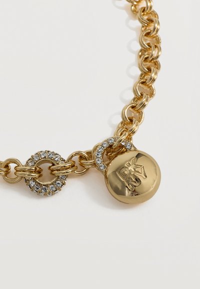 Bracelet chaîne en or avec un pendentif circulaire gravé "DKNY" et des anneaux sertis de cristaux reliant les segments de la chaîne.