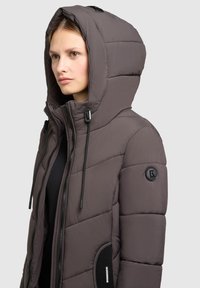 Dunkelgraue gepolsterte Jacke mit Kapuze, ausgestattet mit einem Reißverschluss vorne, Kordelzügen und gerippten Akzenten an den Taschen. Logo am Ärmel.