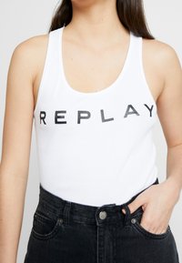 Canotta bianca in cotone con il logo "REPLAY" in bold lettere nere. Presenta un scollo rotondo e un design aderente. Abbinata a jeans neri a vita alta.