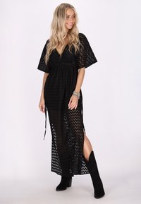 Abito maxi in pizzo nero con maniche corte, scollo a V profondo e spacchi laterali. Presenta una vita cintrata e un tessuto fluido e testurizzato.