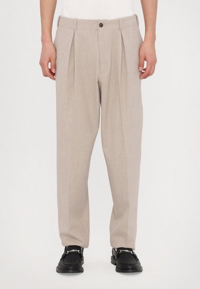 Pantalons plissés beiges en tissu texturé, dotés d'une fermeture à bouton et de passants de ceinture, associés à des chaussures noires.