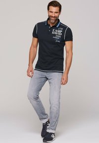 Granatowa koszulka polo z kontrastowym kołnierzykiem, z białym i niebieskim napisem oraz grafiką. Połączona z jasnoszarymi dżinsami i ciemnymi sneakersami.