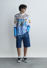 Giovane con indosso una maglietta a maniche lunghe Billionaire Boys Club con stampa grafica, pantaloncini di jeans, sneaker nere e bianche e occhiali da sole, in piedi contro un muro semplice.