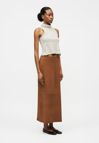 Γυναίκα που στέκεται φορώντας ένα αμάνικο ριγέ crop top, μακρύ καφέ φούστα και μαύρα παπούτσια μπαλαρίνες, μπροστά σε ένα απλό φωτεινό φόντο.