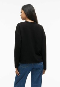 Juodas ilgomis rankovėmis marškinėliai su laisvu kirpimu, pasižymintys nusileidusia pečių linija ir suapvalintu iškirpimu. Suderinti su mėlynomis aukštai pasaitinėmis džinsomis.