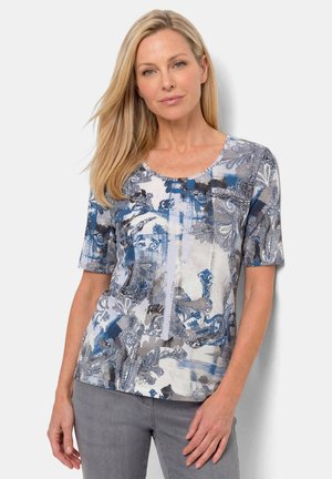 Frau mit glatten blonden Haaren, die eine kurzärmelige Bluse mit blauem, grauem und weißem Muster und graue Hose trägt, steht vor einem weißen Hintergrund.