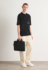 Tricou din bumbac negru cu buzunar pe piept și logo-ul "HUGO", asortat cu pantaloni de culoare crem și o geantă pentru laptop de culoare neagră, cu fermoar. Adidași albi.
