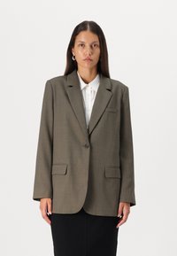 Blazer vert olive avec un seul bouton, revers crantés, deux poches à l'avant et épaules structurées. Porté par-dessus une chemise blanche à col.