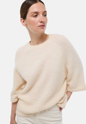 Femme aux cheveux bruns tirés en arrière, portant un doux pull en crème duveteux avec des manches jusqu'aux coudes et un pantalon crème, regardant sur le côté.