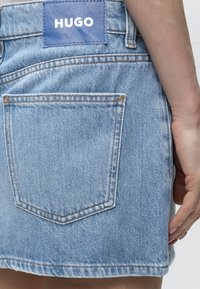 Φούστα από ανοιχτό μπλε denim με μία πίσω τσέπη και μπλε ετικέτα με την ένδειξη HUGO στη μέση, χέρι που ακουμπά δίπλα στη φούστα.