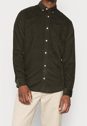 Homme portant une chemise en velours côtelé vert foncé à boutons avec poche poitrine et un pantalon beige, debout devant un fond uni.