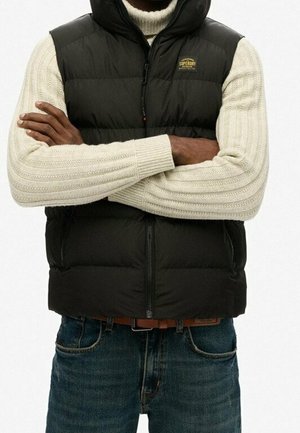 Mężczyzna w czarnej pikowanej kamizelce na kremowym swetrze w warkocze, z założonymi rękami, w połączeniu z niebieskimi dżinsami i brązowym paskiem.