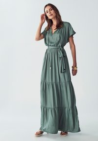 Maxi šaty z jemné zelené látky, s krátkými rukávy, páskem v oblasti pasu a vrstveným designem s volným střihem. Minimalistické detaily a hladká textura.