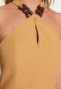 Halterneckklänning i senapsfärgad tyg med ett nyckelhålskärning vid bröstet och en mörkbrun kedjeaccent vid halsringningen.