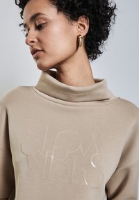 Beige sweatshirt med hög krage och glansig, upphöjd text "SLOW VIBES". Modellen har kort, lockigt hår och en guldring i örat.