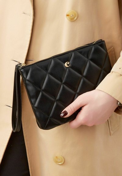 Clutch in pelle nera trapuntata con hardware dorato e una tracolla staccabile, tenuto contro un cappotto beige con un grande bottone.