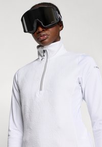 Camisola de fleece branca com fecho, com padrão texturizado de folhas, apresentando um colar alto e pescoço com fecho. Óculos de esqui pretos com lentes espelhadas.