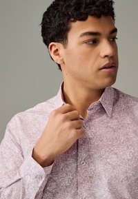 Camicia rosa chiaro con bottoni e motivo dettagliato di case, dotata di colletto alla coreana e maniche lunghe. Mani che sistemano il colletto.