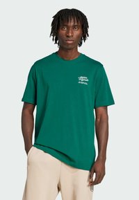 Green cotton t-shirt with a round neckline and short sleeves, featuring white logo text on the left chest area. Casual fit.