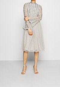 Robe gris clair avec des manches longues ornées, des poignets évasés et une jupe en tulle jusqu'aux genoux. Associée à des talons roses pâles à lanières.