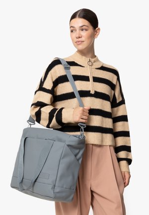 Borsa tote grigia con superficie texturizzata, forma rettangolare, doppie maniglie e tracolla regolabile. Indossata con un maglione a righe beige e nere.